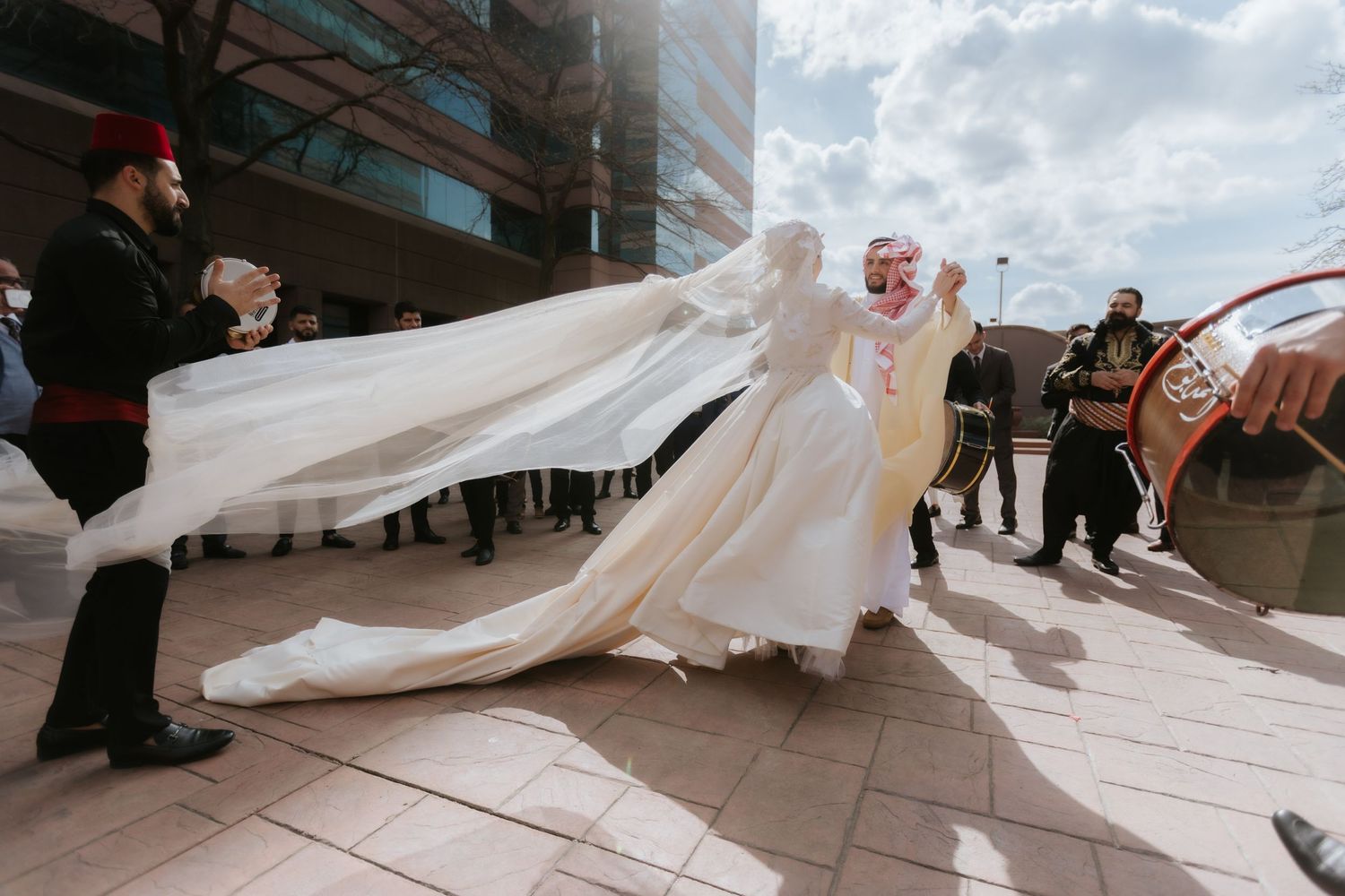 Bride in white gown with flowing veil during outdoor wedding celebration with guests in traditional attire.