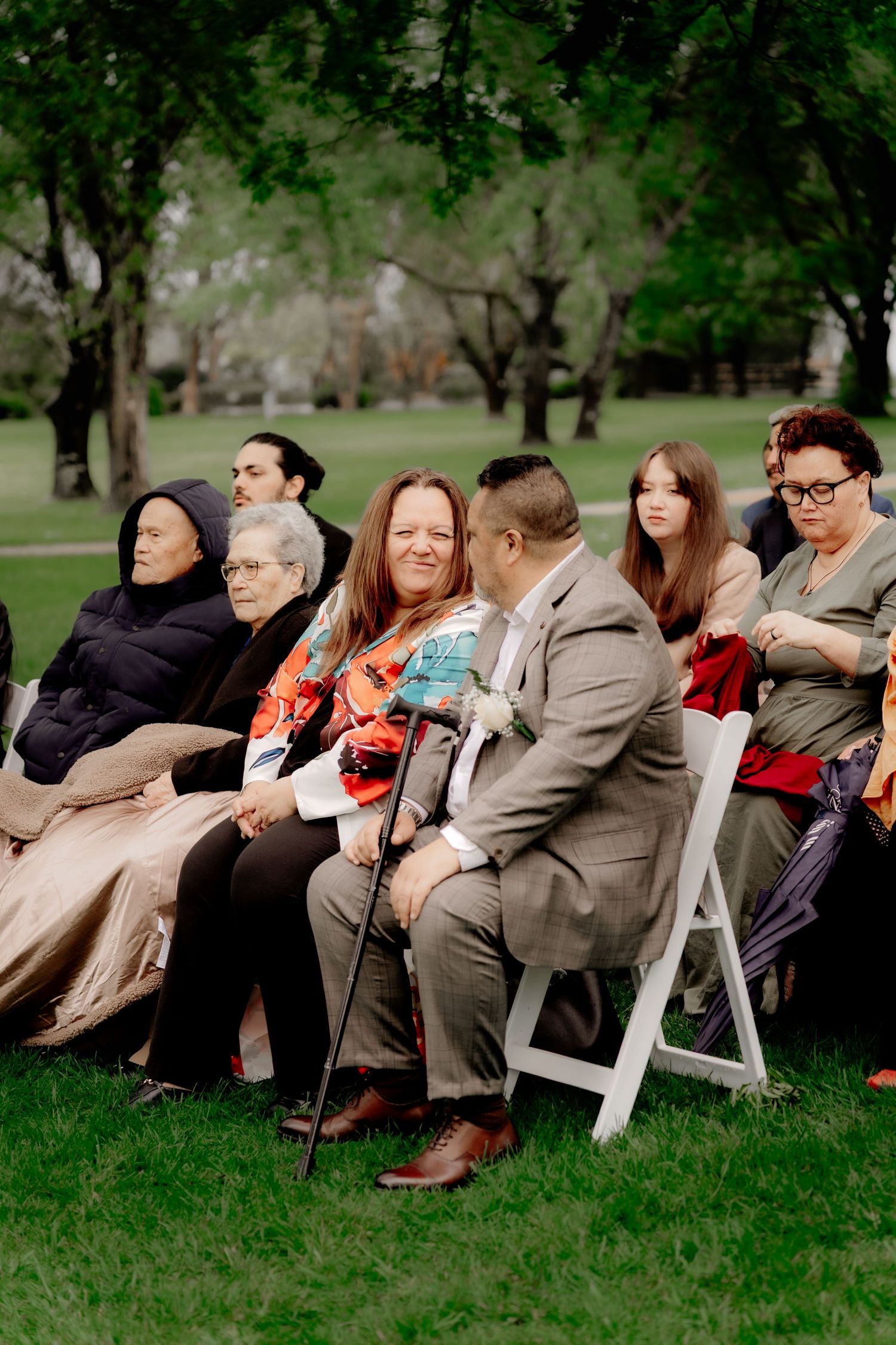 Guests seated on white chairs at an outdoor wedding ceremony on a green lawn surrounded by trees.