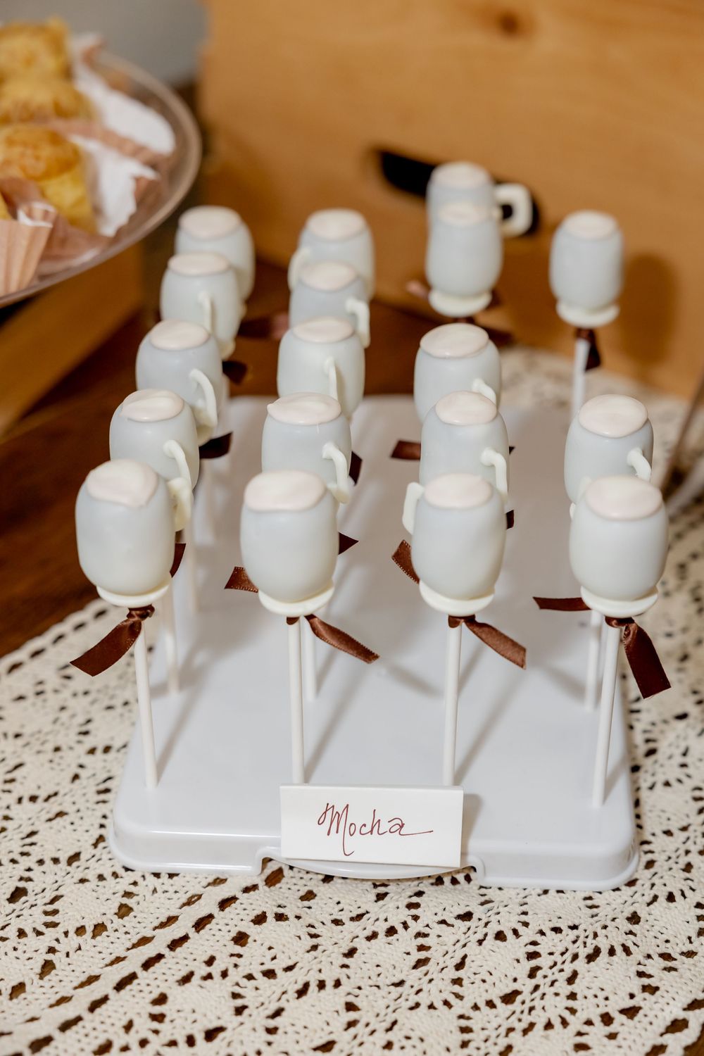 White mocha cake pops shaped like coffee mugs on a display stand with brown ribbon bows and a label card.