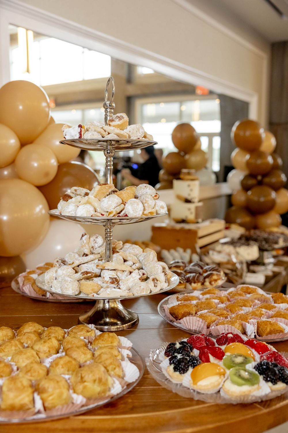 Elegant bridal shower dessert table with tiered pastry stand, cream puffs, fruit tarts, and gold balloon decorations at a party.