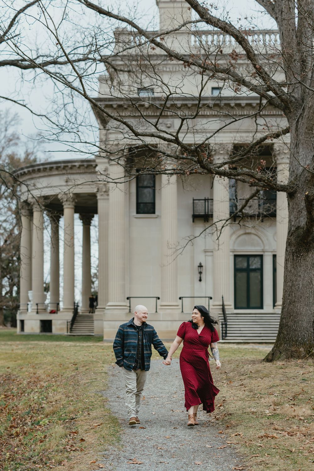 Couple holding hands walking path before grand white colonial mansion with columns in winter.