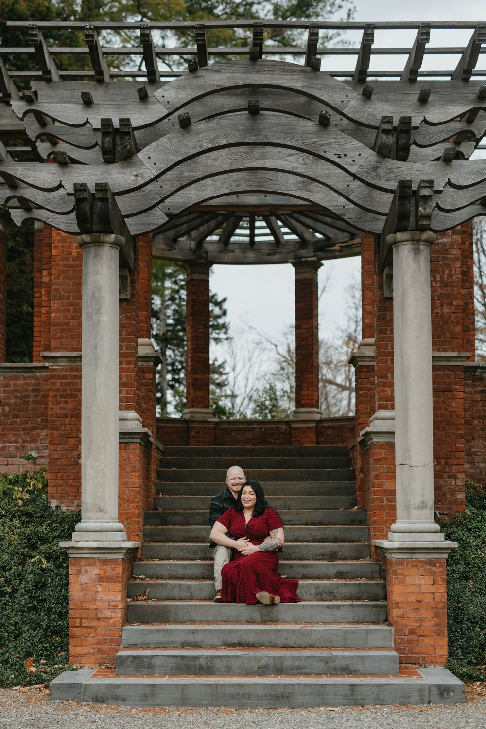 Couple sits on stone steps under elegant pergola with brick columns, woman in red dress embraced by partner in garden setting.