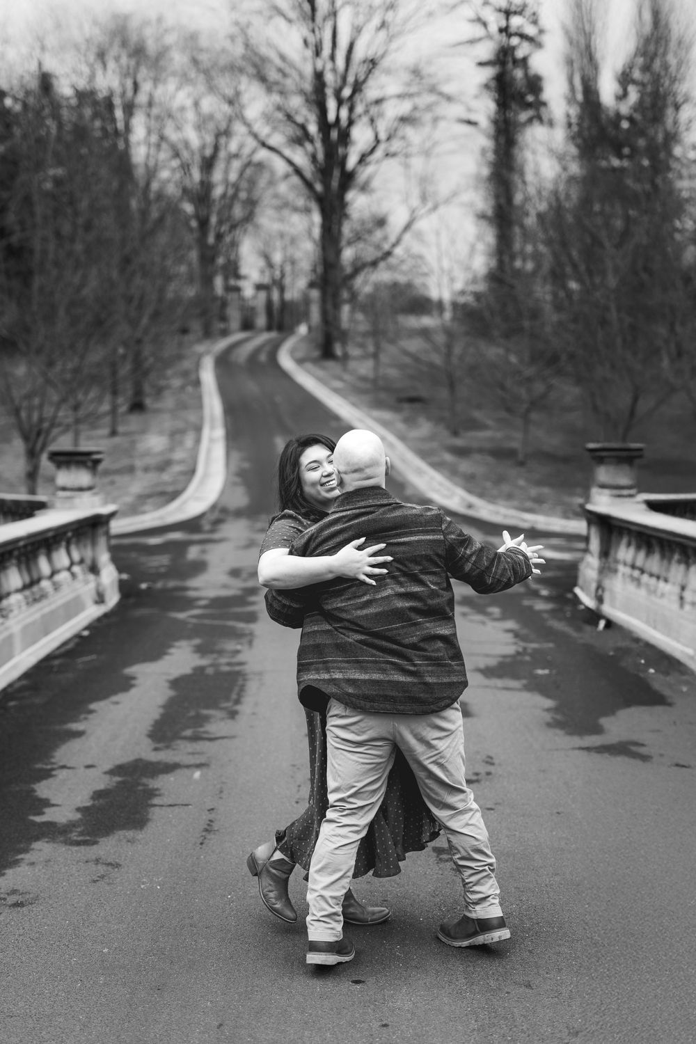 Black and white photo of a couple embracing on a bridge in a park during winter.