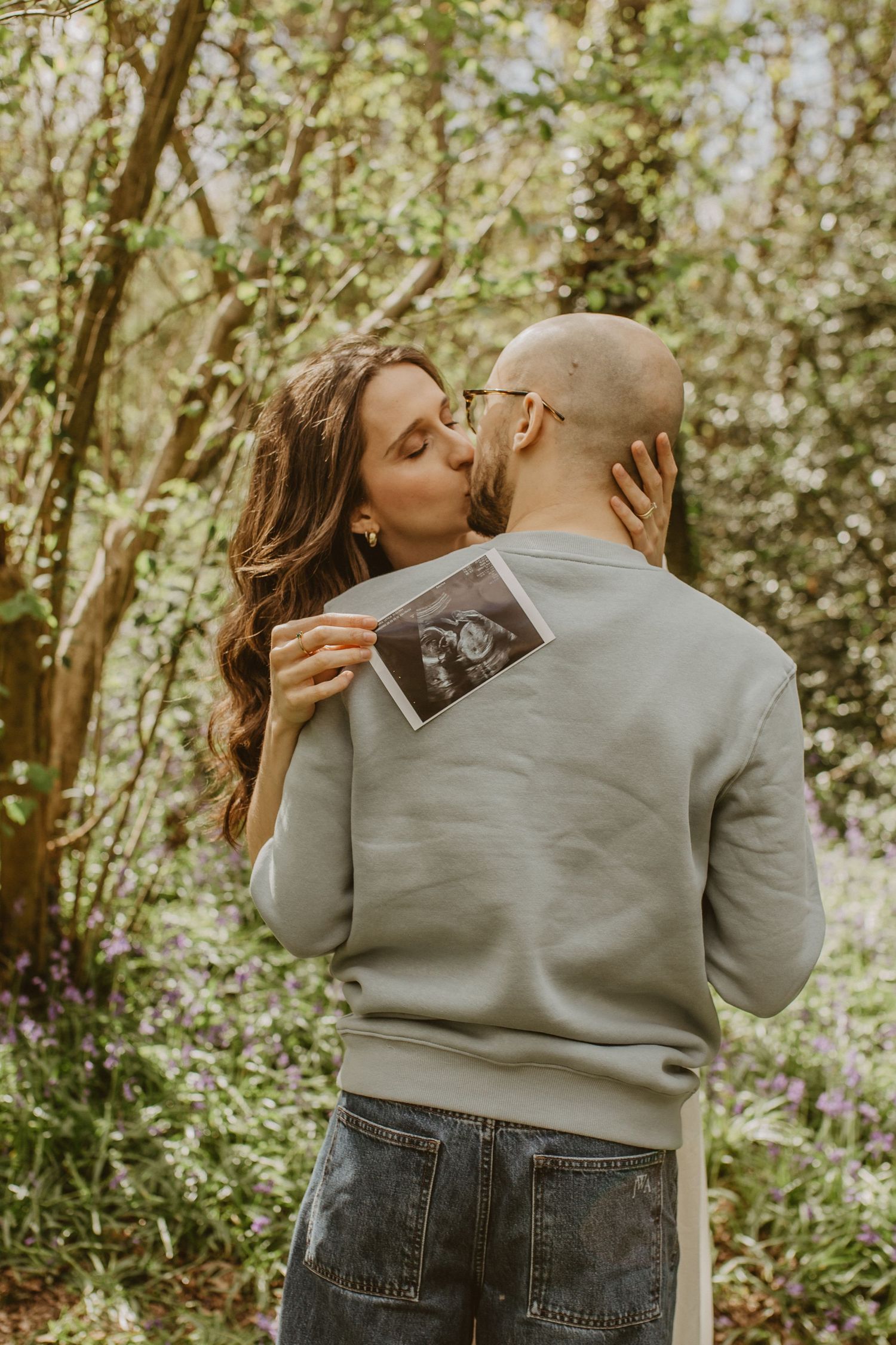 Couple kissing in woodland setting while holding ultrasound scan photo, announcing pregnancy outdoors.