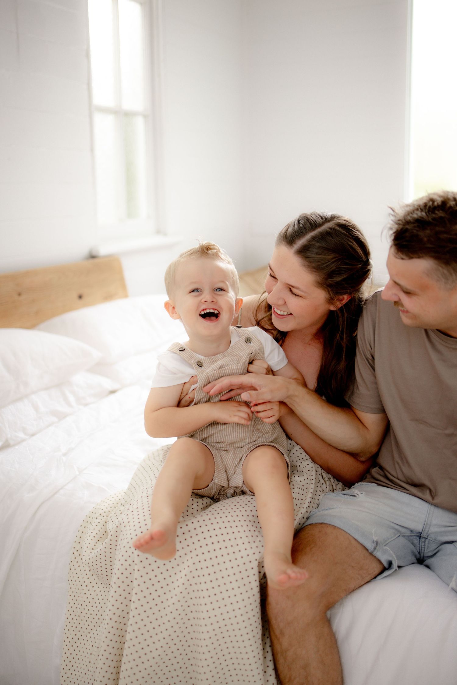 Happy family with laughing toddler sitting on white bed, parents smiling and bonding in bright bedroom.