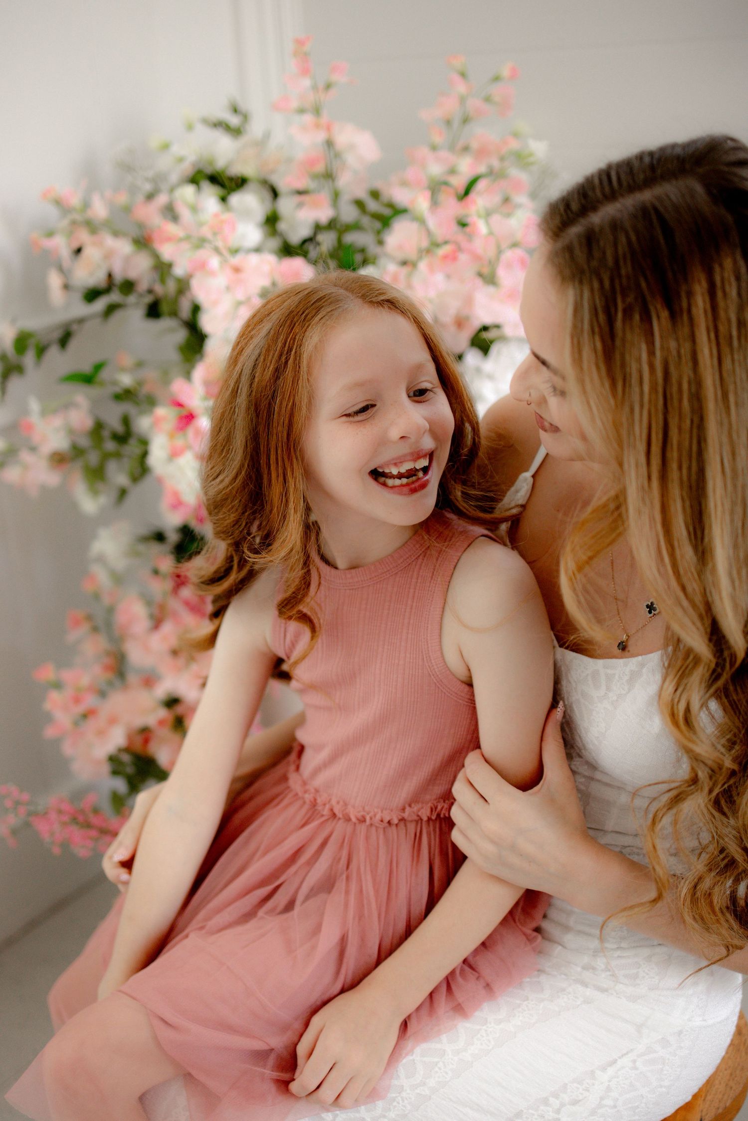 A joyful mother and daughter in pink dresses laugh together in front of a beautiful floral backdrop.