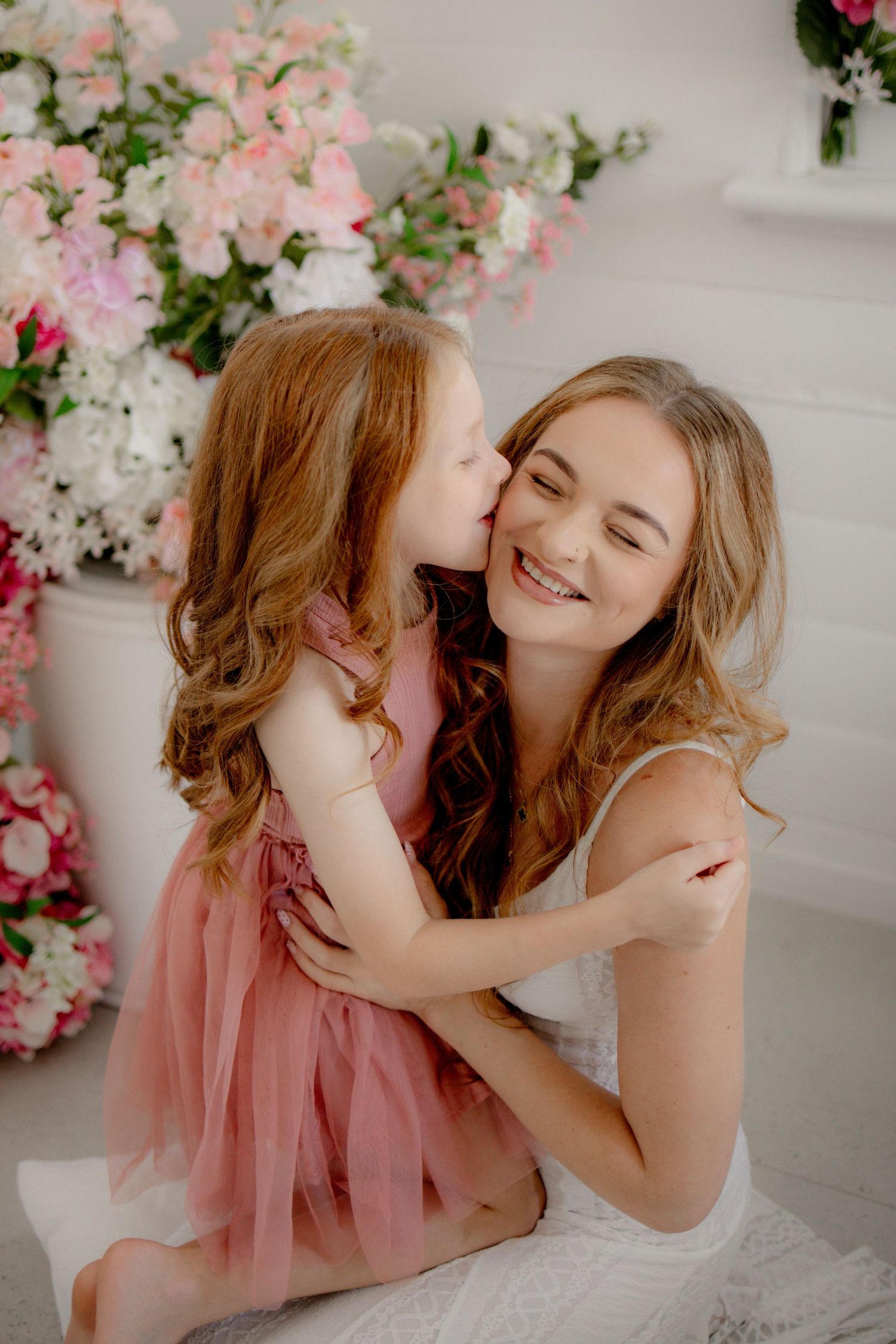 A smiling mother in white dress receives a kiss from her young redhead daughter in a pink tutu dress, surrounded by flowers.