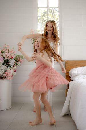 A joyful mother twirls her redhead daughter in a pink tutu dress in a bright white bedroom decorated with pink flowers.
