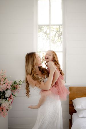 A joyful mother in a white dress lifts her laughing little girl in a pink tutu in a bright, flower-filled white room.