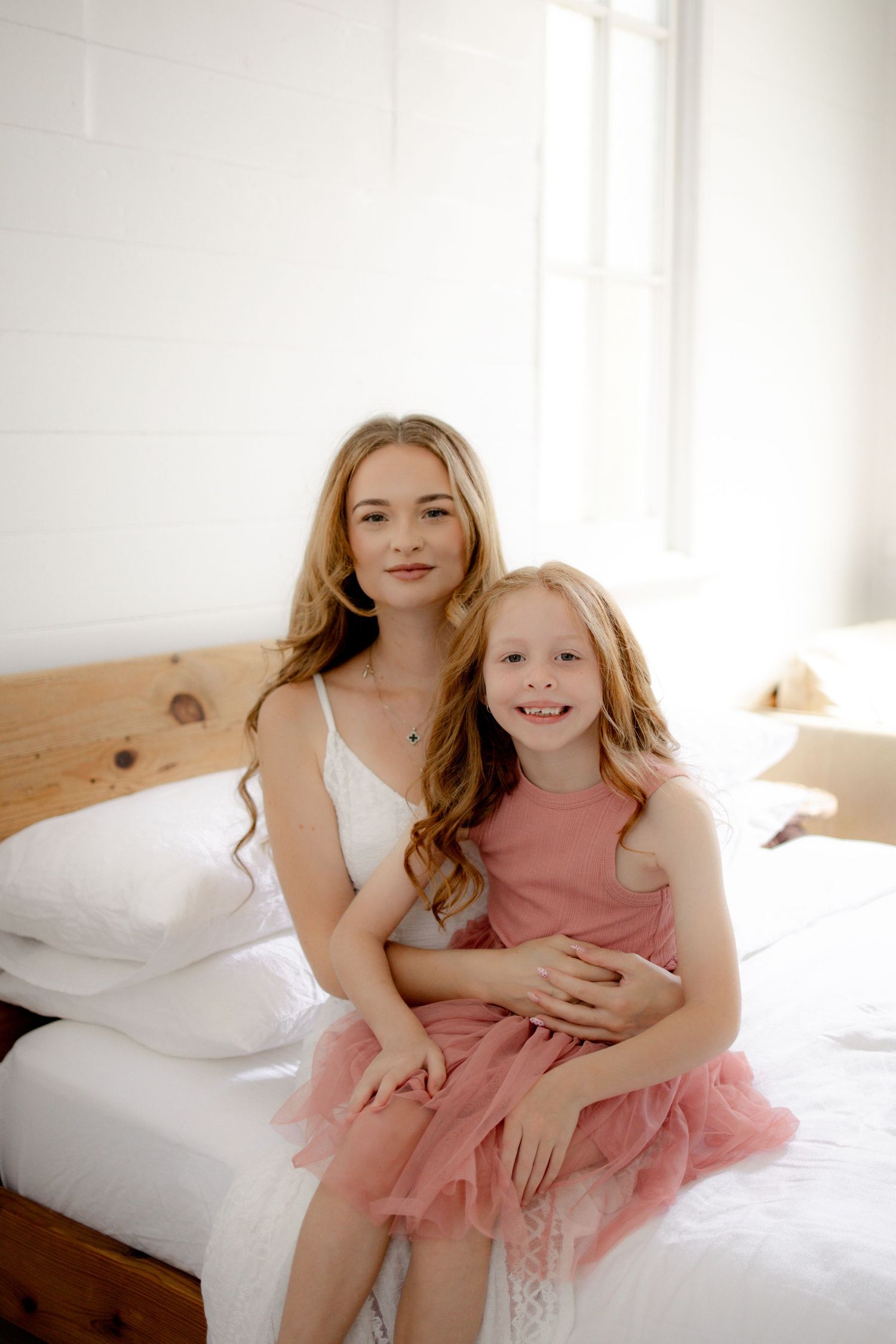 A young woman in a white dress sits on a bed hugging a smiling little girl in a pink tulle dress.