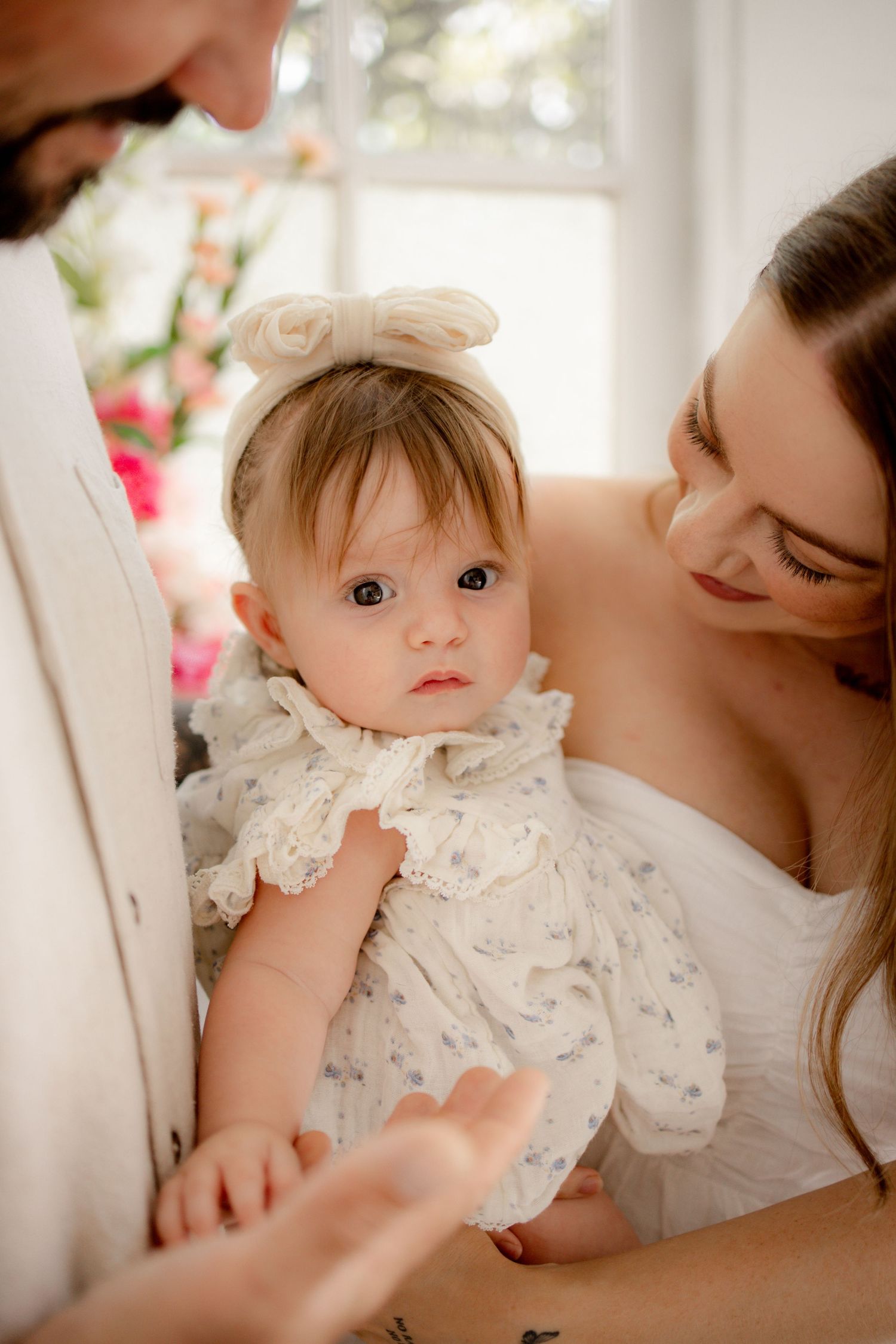 A baby girl in a white floral dress and bow headband is held by her parents during a bright, elegant family portrait.