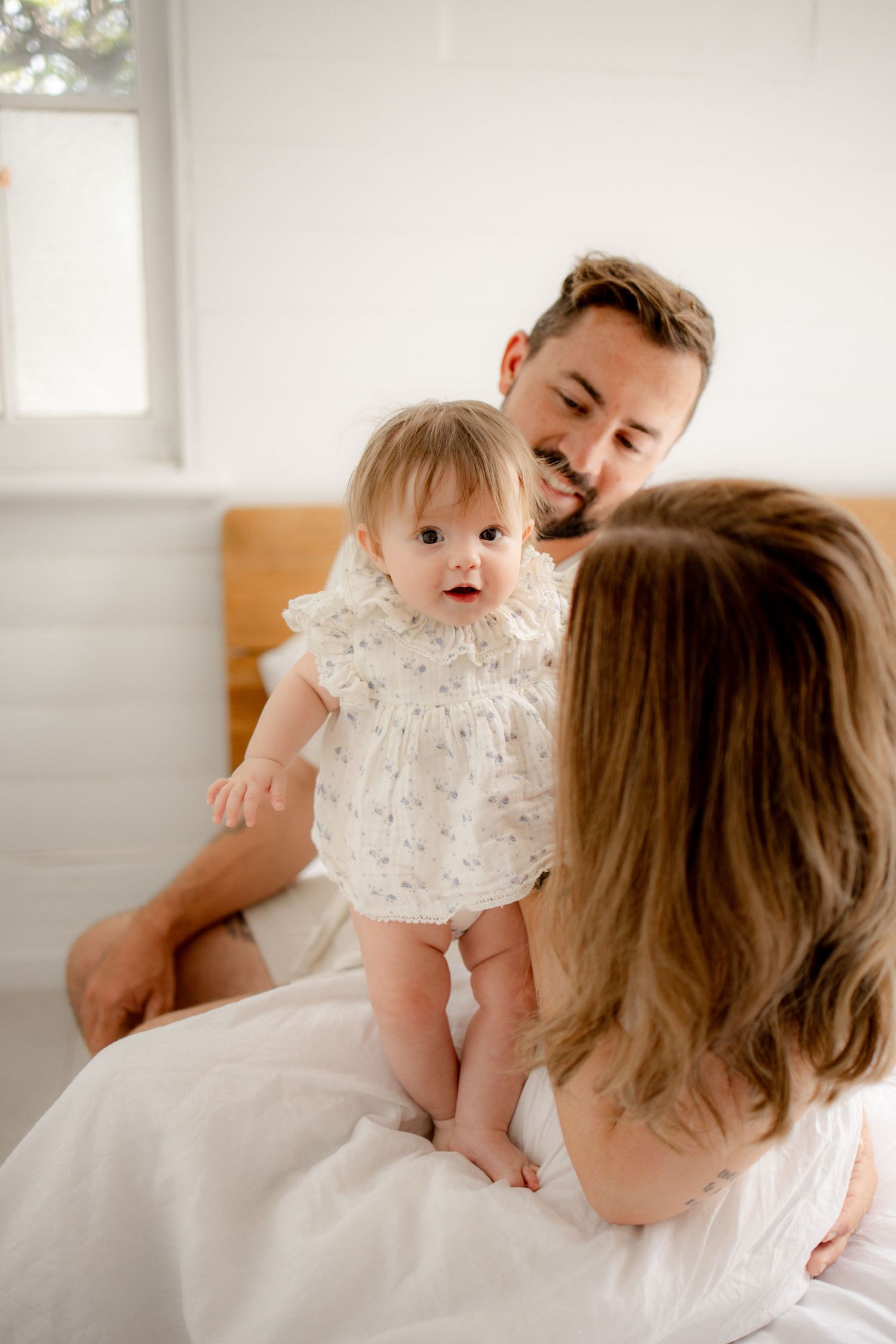 A smiling father and mother interact with their baby girl in a white floral romper on a bright, airy bed.