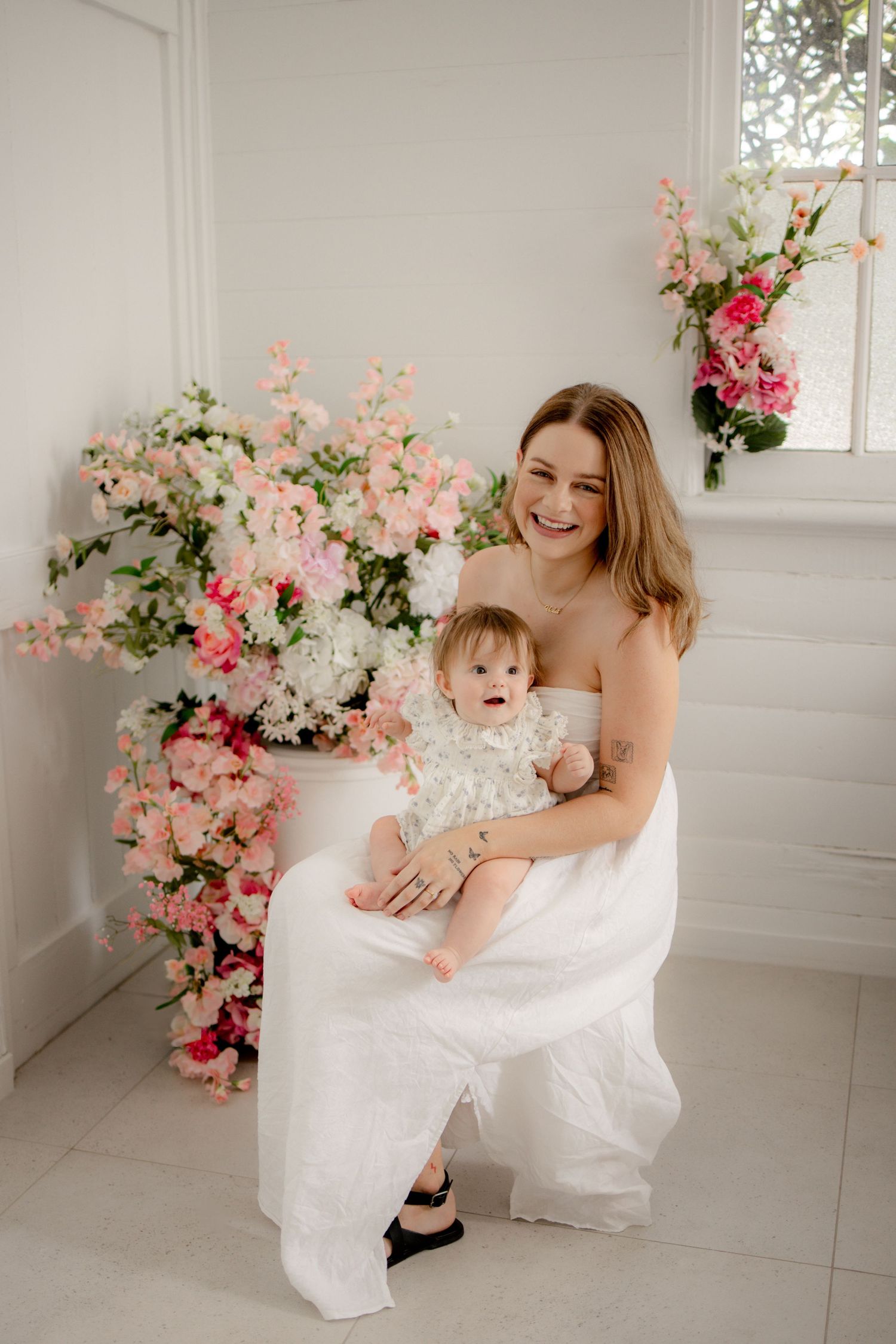 A smiling mother in white holds her baby girl amid lush pink and white floral arrangements in a bright studio.