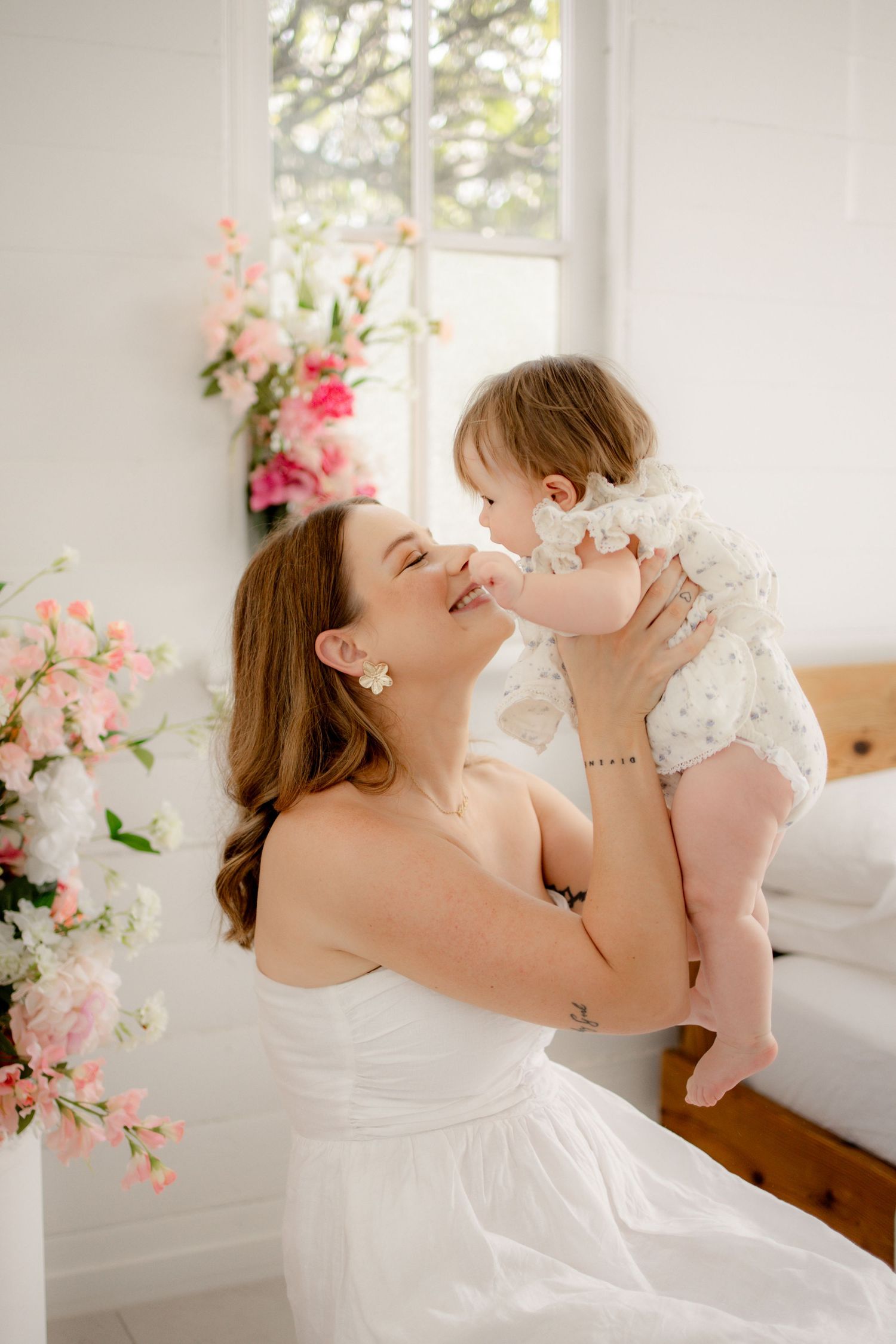 A mother in a white dress lovingly holds her baby girl near pink flowers in a bright, airy room.
