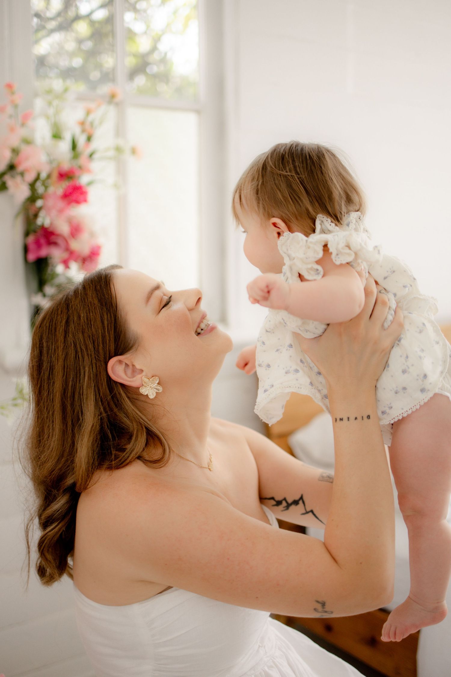 A smiling mother in a white dress lifts her baby girl in a bright, floral-decorated room.