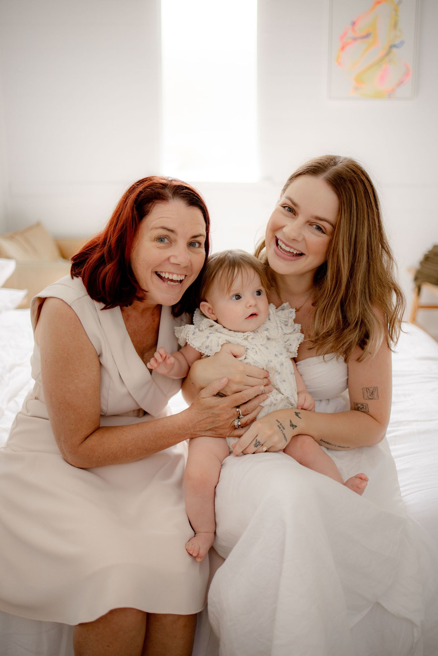 Two women and a baby smiling together on a white bed in a bright bedroom, dressed in white outfits.