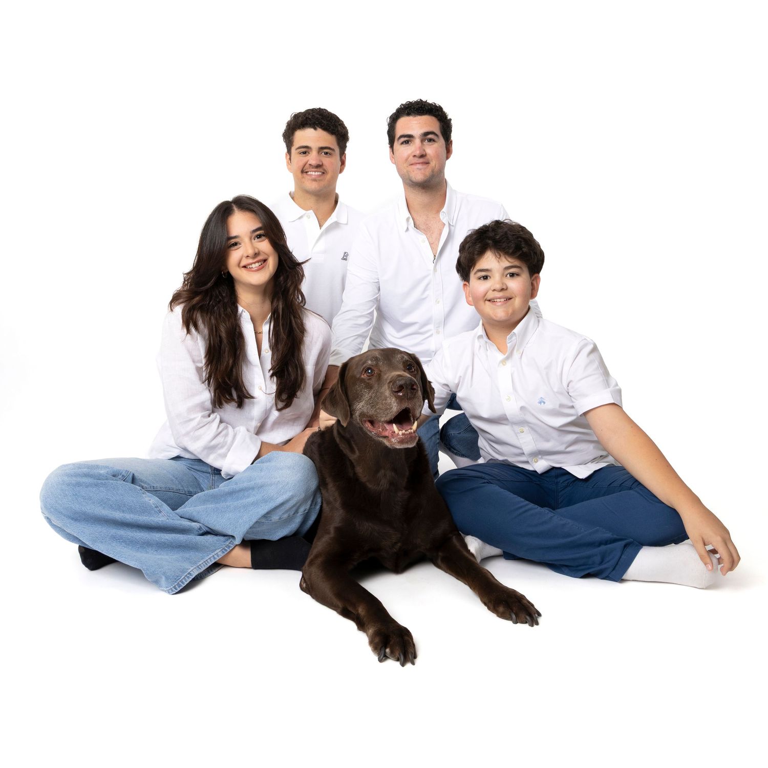 Family of four in white shirts posing with a chocolate Labrador on a white background.