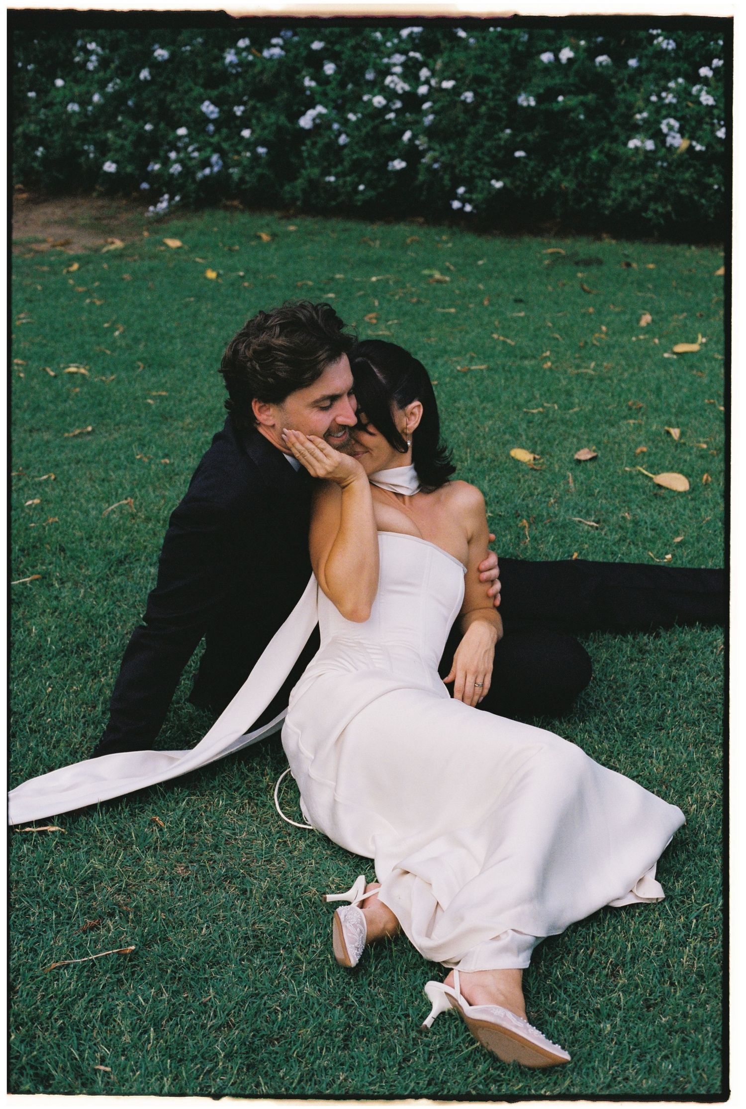 Bride and groom sitting on green grass, sharing a romantic moment, bride in white strapless gown, groom in black suit.