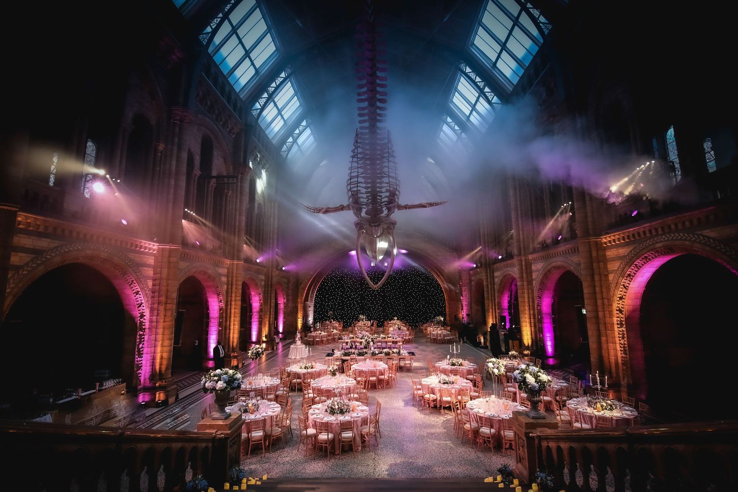 Elegant wedding reception in a grand hall with pink uplighting, round tables, and a dramatic whale skeleton hanging overhead.