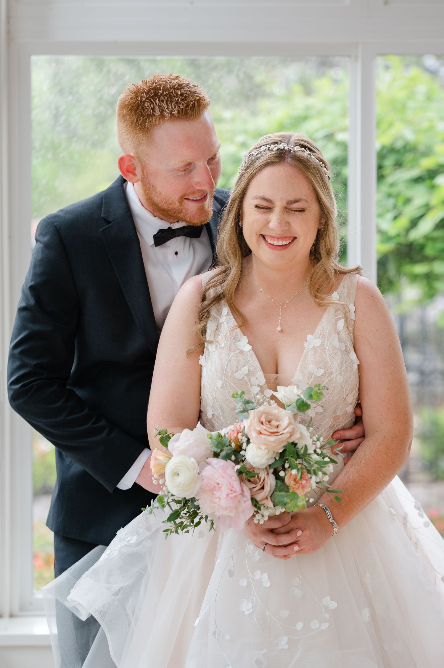 Smiling bride in floral gown holds pink bouquet as groom in black tuxedo embraces her near bright window.