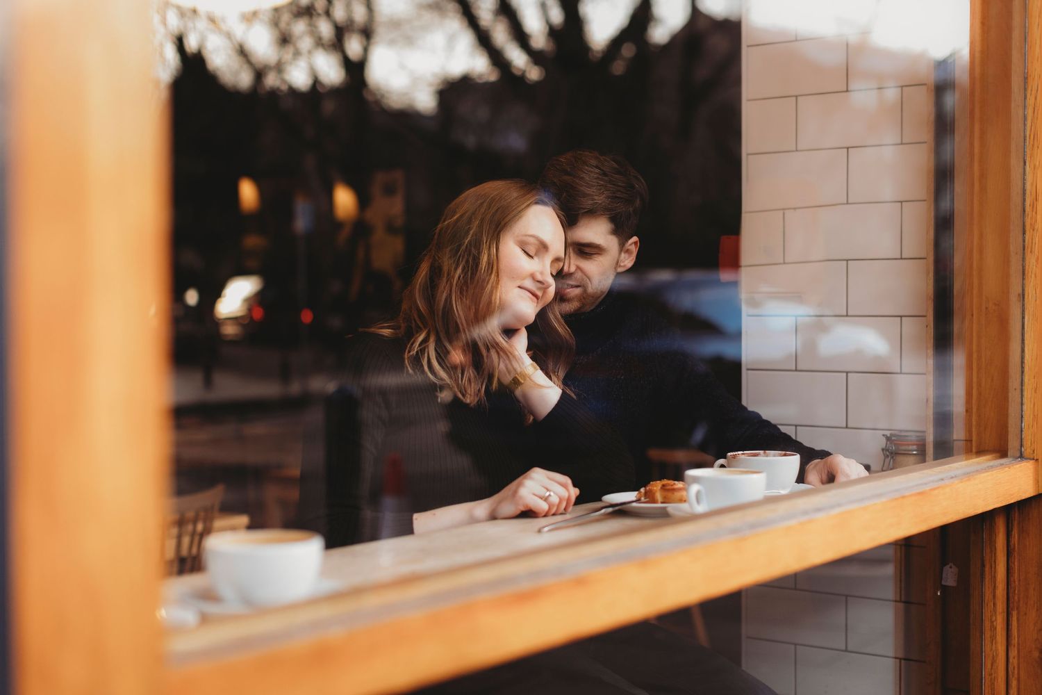 Couple sitting together inside a cafe, viewed through a window with reflections of historic buildings and a church tower.