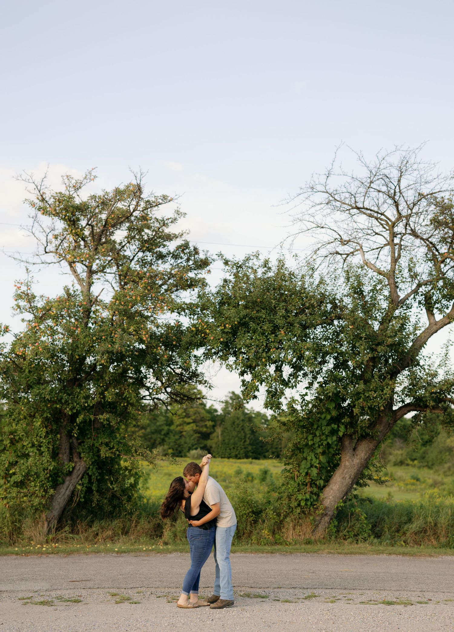 Couple sharing a romantic embrace in a parking lot surrounded by lush green trees and open fields at dusk.