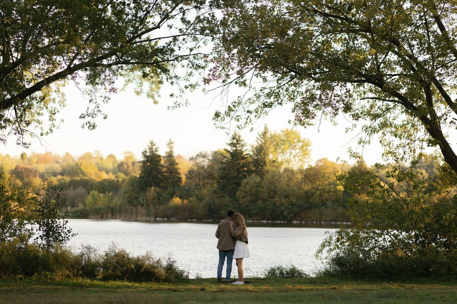 Couple standing together by a tranquil lake surrounded by autumn trees in a romantic outdoor setting.