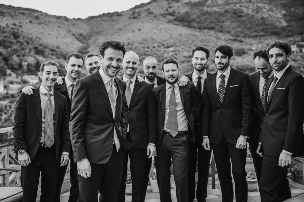 Groomsmen in dark suits pose together for a black and white group photo outdoors.