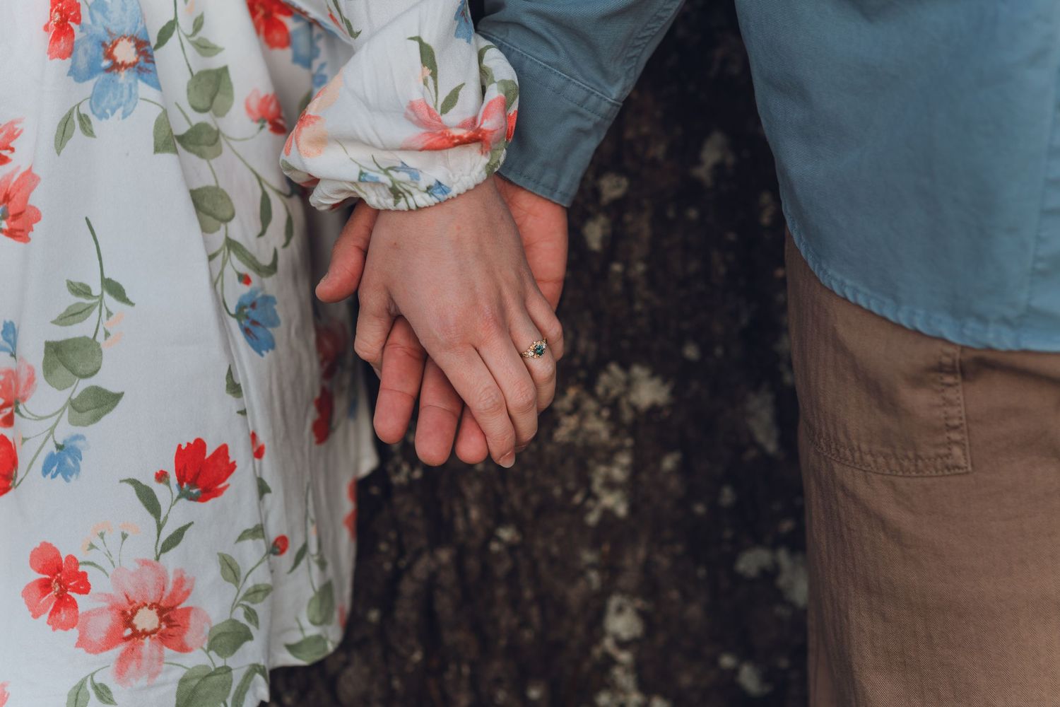 Close-up of a couple holding hands, woman wearing a floral dress with an engagement ring visible.