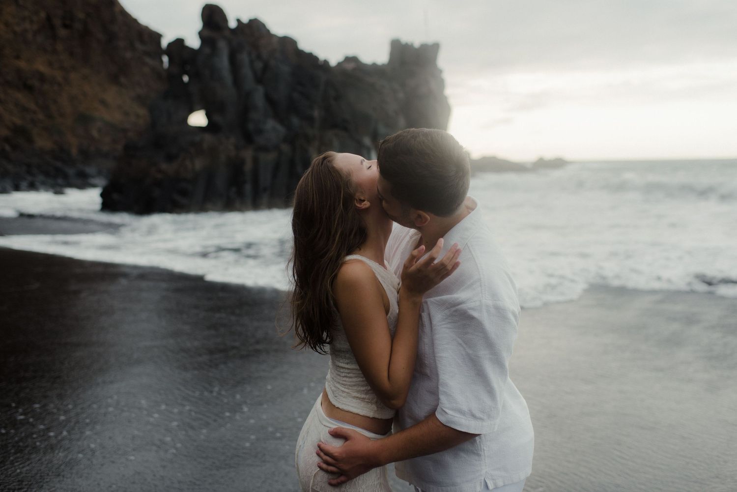 Couple kissing on black sand beach surrounded by colorful floral arrangement with dramatic cliffs and ocean waves behind them.