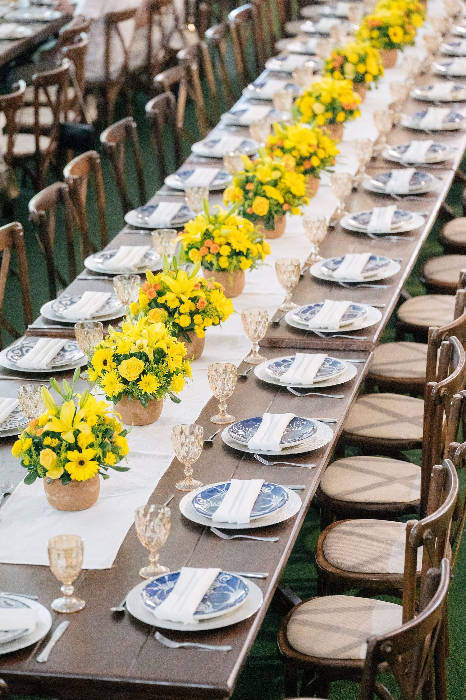 Elegant outdoor banquet table with yellow floral centerpieces, blue and white plates, crystal glasses, and wooden cross-back chairs.
