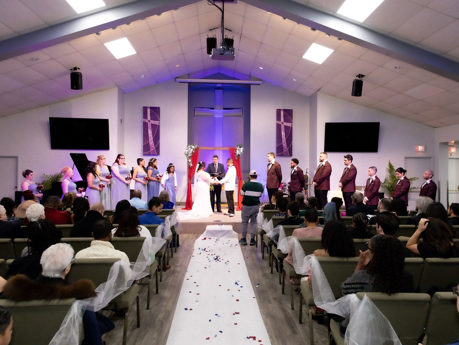 Wedding ceremony in a church with bridal party, white petal aisle, and guests seated in decorated pews.