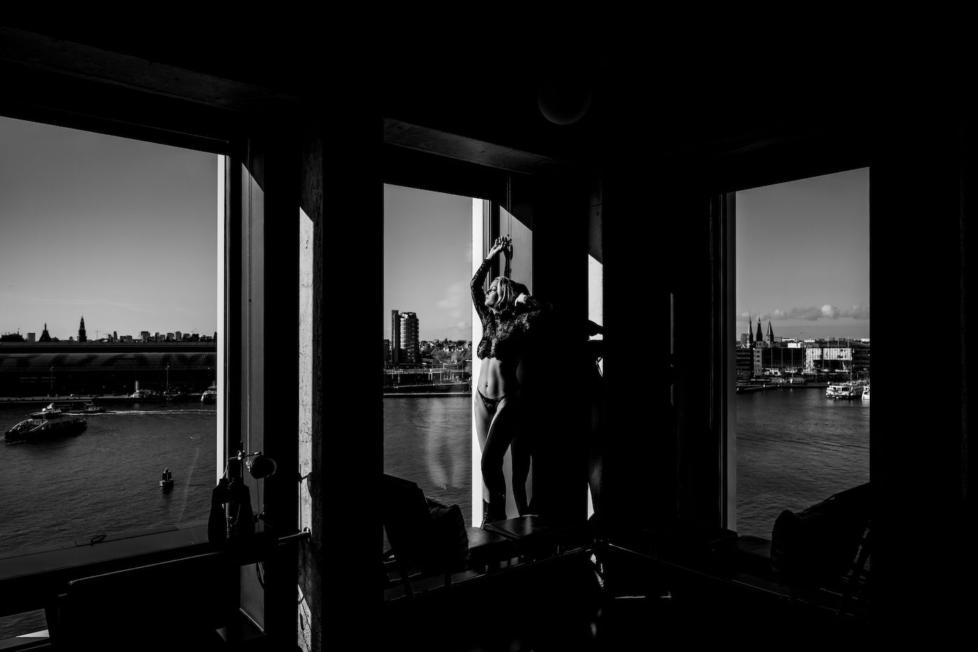 Silhouetted person standing by large windows overlooking a river and city skyline in black and white.