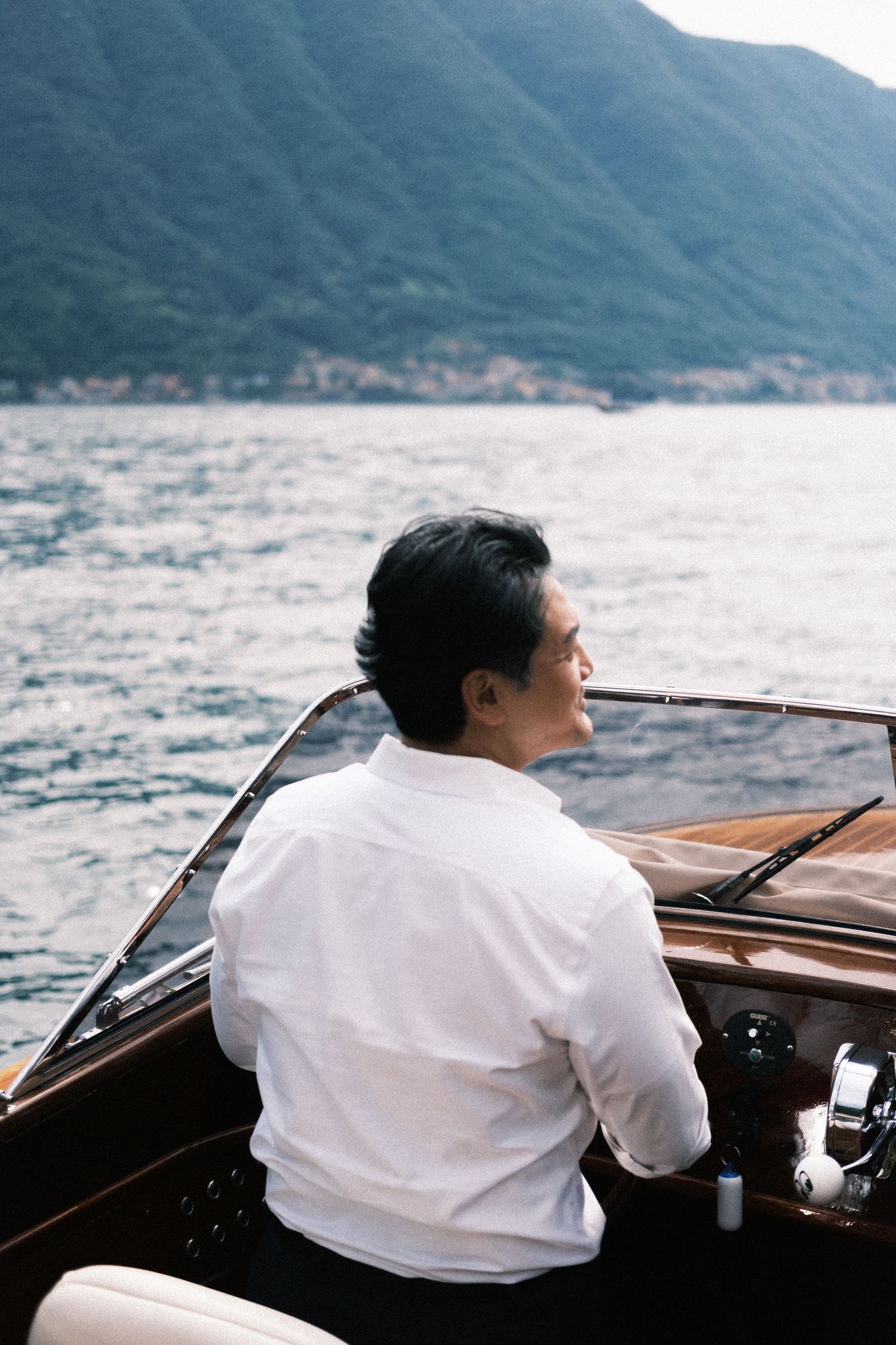 Man in white shirt sitting in a luxury wooden speedboat on a calm lake with mountain backdrop.