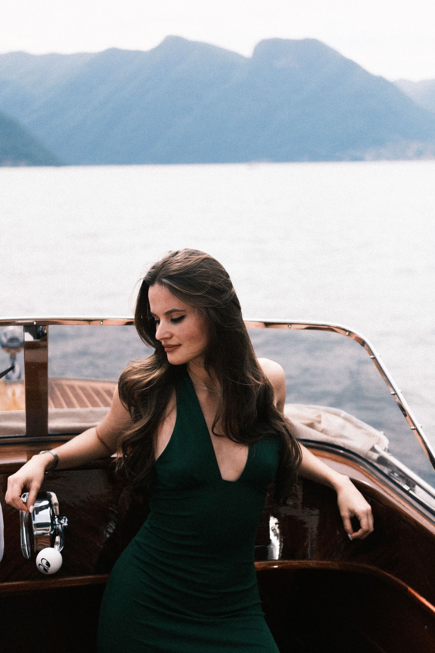 A woman in an elegant green halter dress sits on a wooden boat with mountain lake views in the background.