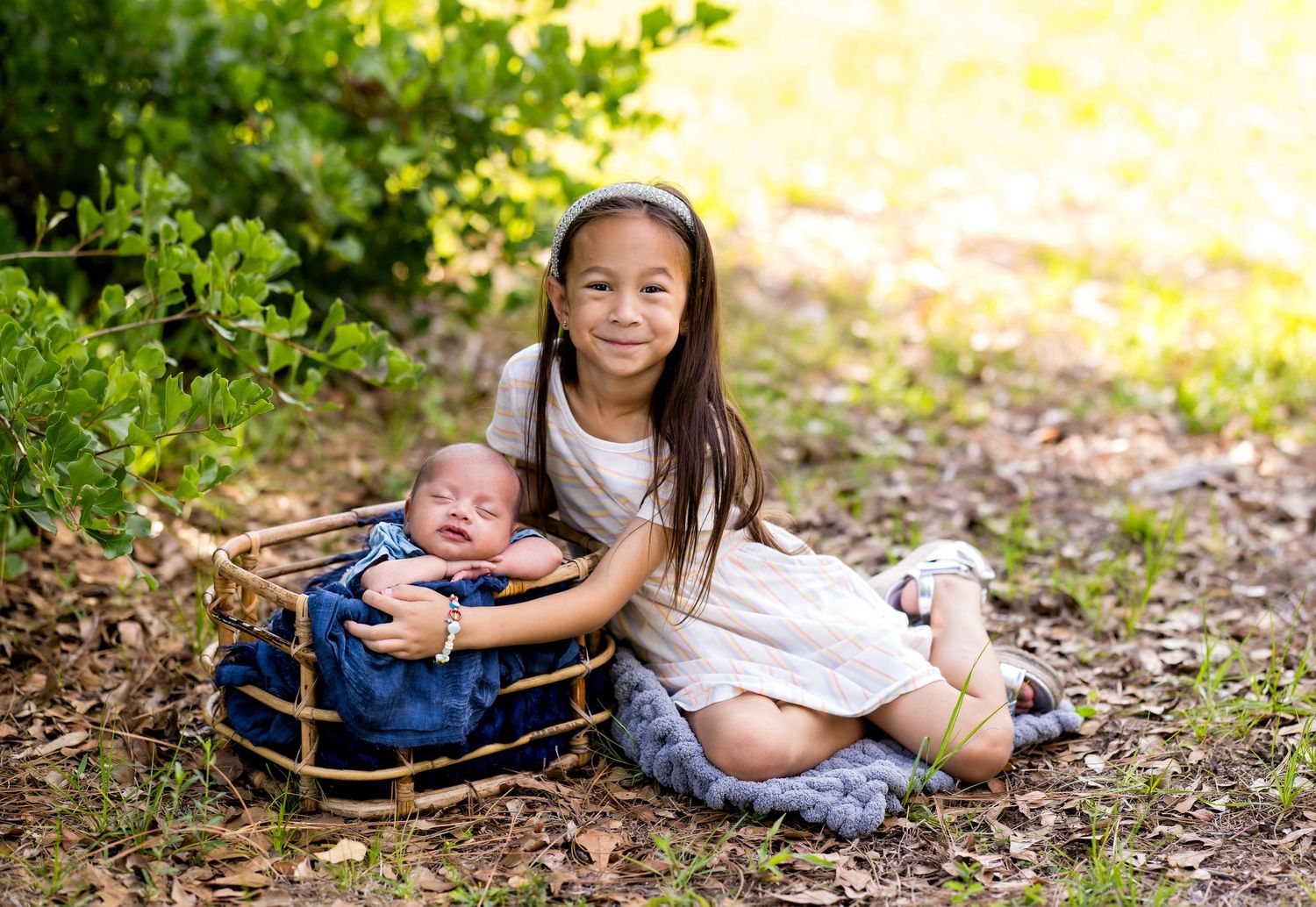 Young girl in white dress poses outdoors with sleeping newborn baby in a wicker basket surrounded by greenery.