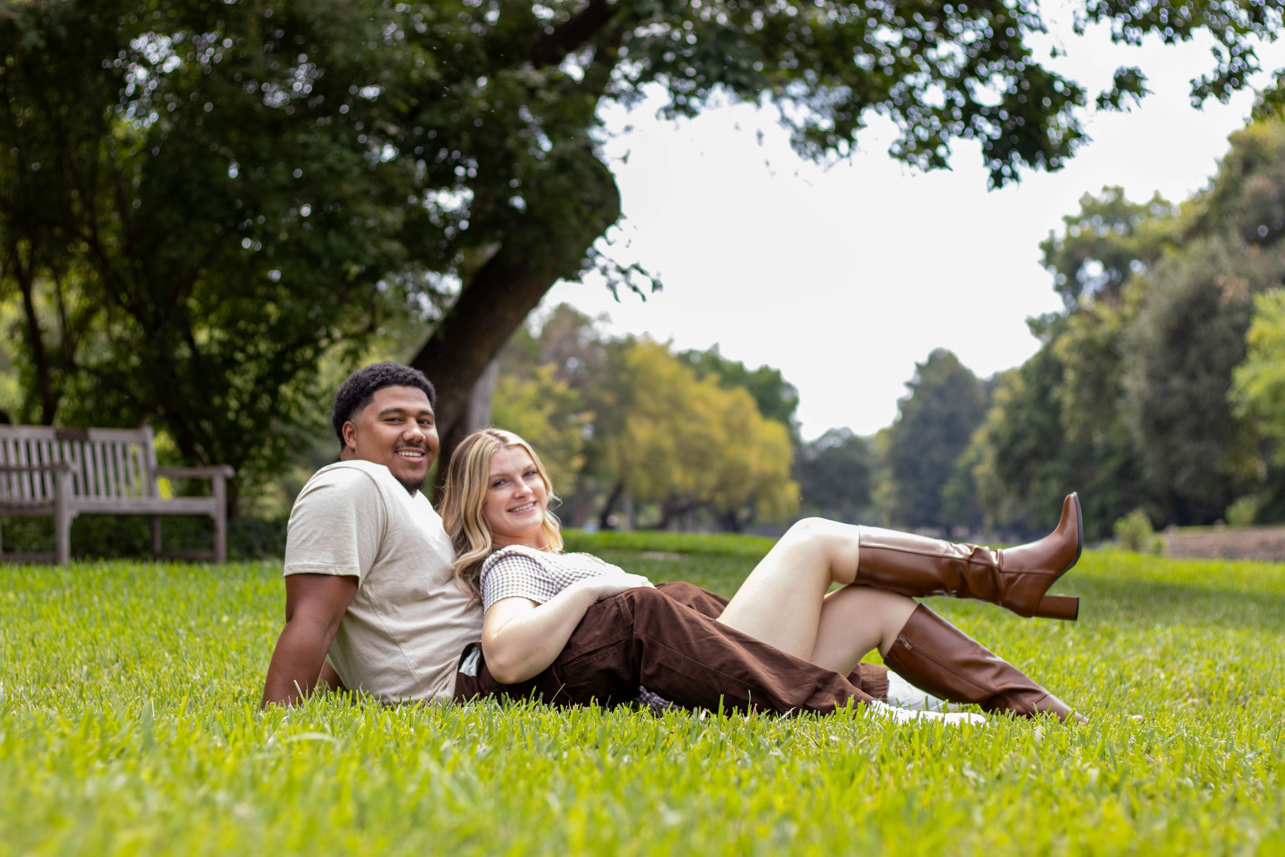 Happy interracial couple sitting together on green grass in a sunny park, smiling and relaxed beneath a large tree.