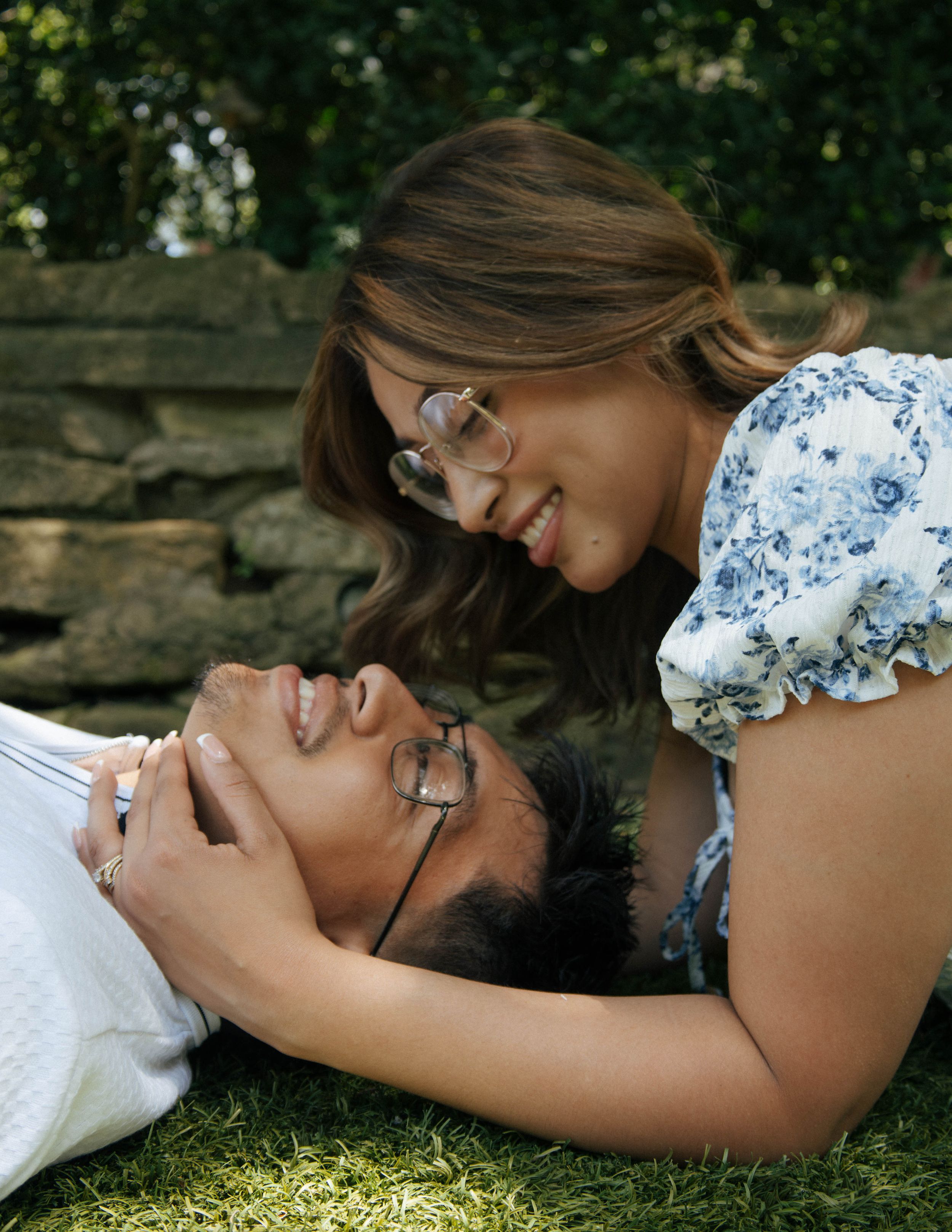 Couple sharing a romantic moment on grass, woman smiling down at man lying in her lap, both wearing glasses.