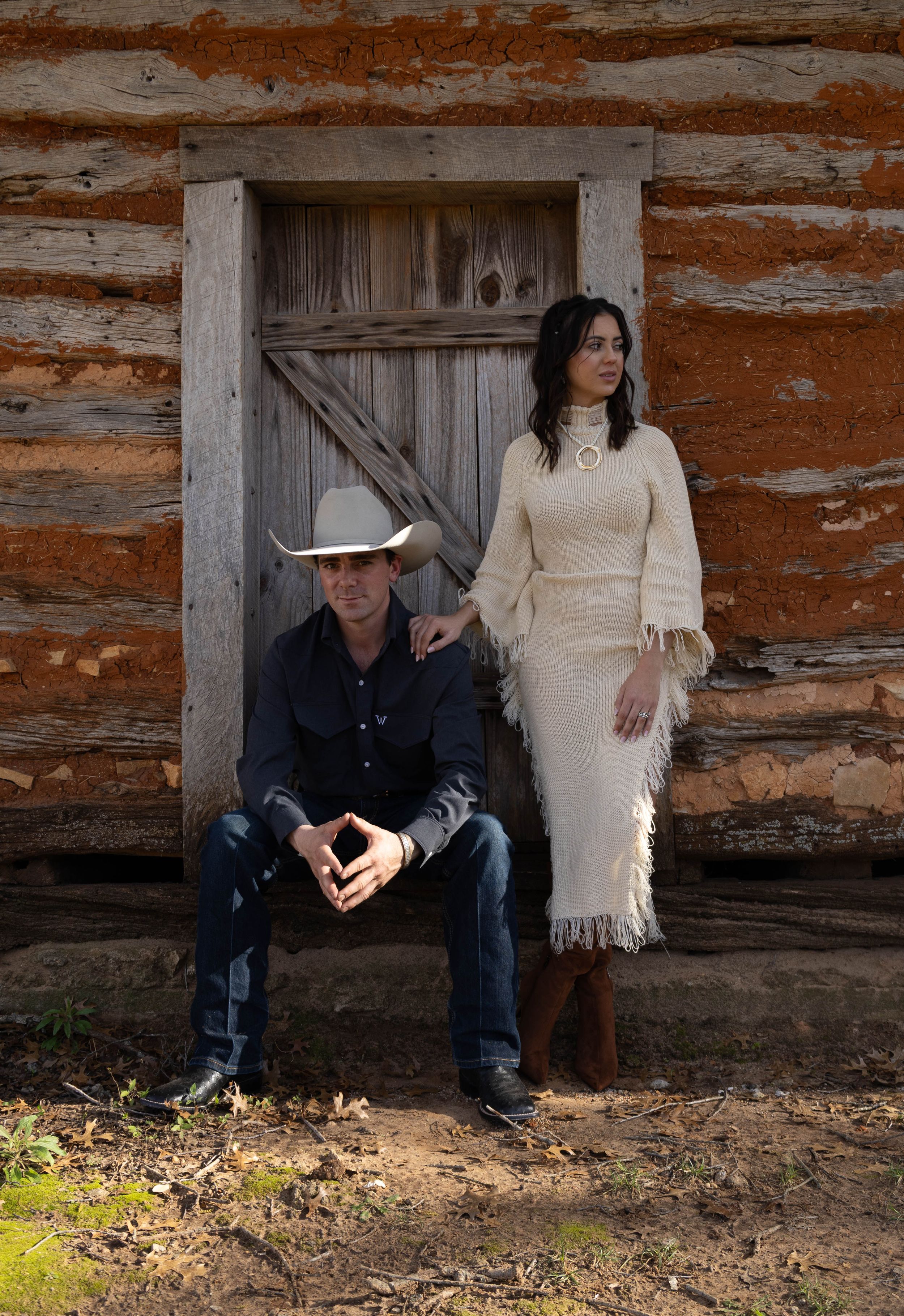 A man in a cowboy hat sits before a rustic log cabin door while a woman in a fringed cream dress stands beside him.