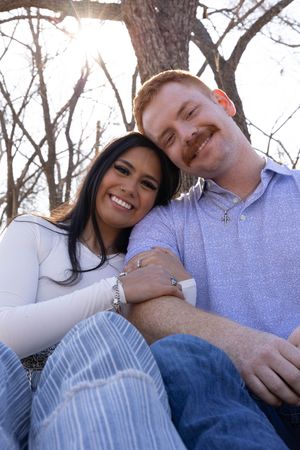 A smiling couple sits together outdoors under a tree, the woman leaning on the man's shoulder in a warm embrace.