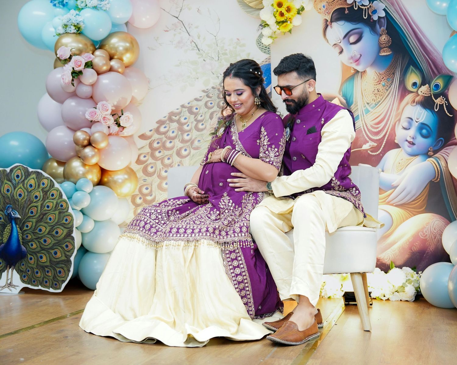 Pregnant Indian couple in traditional attire at a Krishna-themed baby shower with balloon decorations.