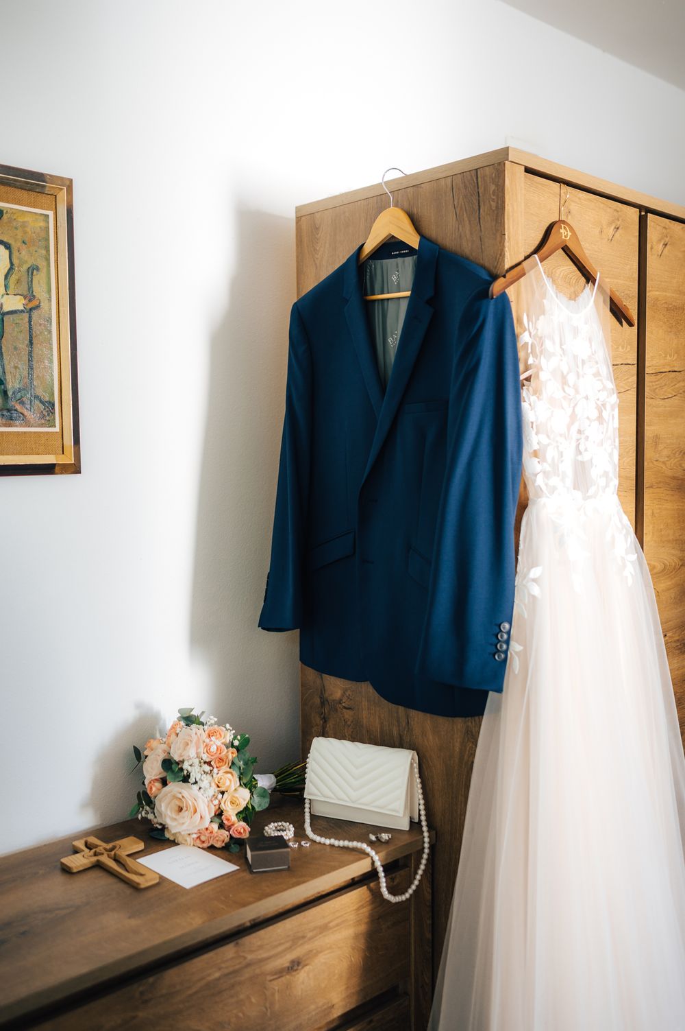 Navy groom suit and lace wedding dress hanging on wooden wardrobe with bridal bouquet and pearl accessories below.