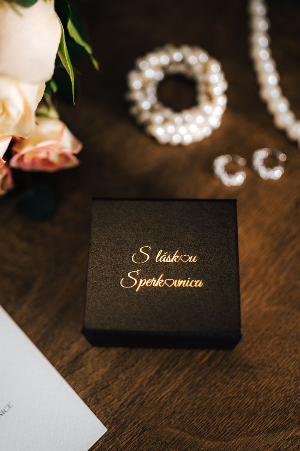 Black gift box with gold 'S láskou Šperkovnica' text beside pearl jewelry and roses on wooden table.
