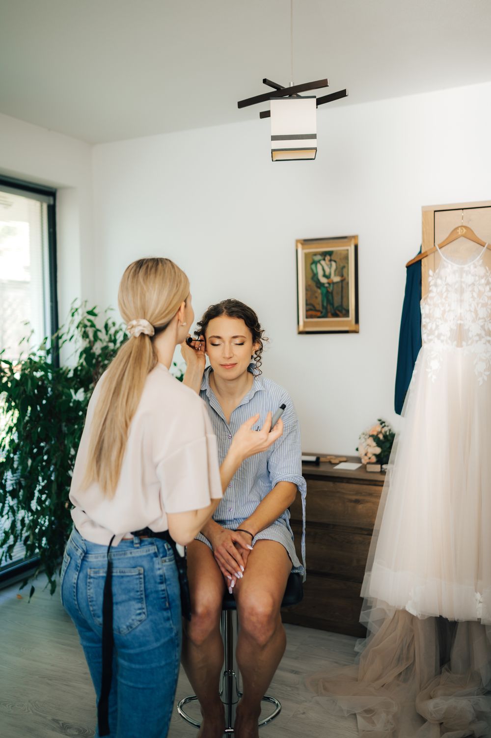 Makeup artist applying makeup to a bride in a bright room with a wedding dress hanging nearby.