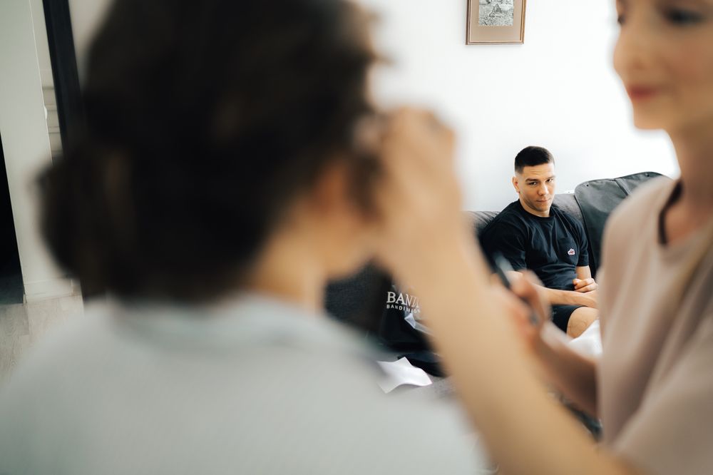 Man in black shirt sits on sofa watching a woman apply makeup to another person in a bright room.