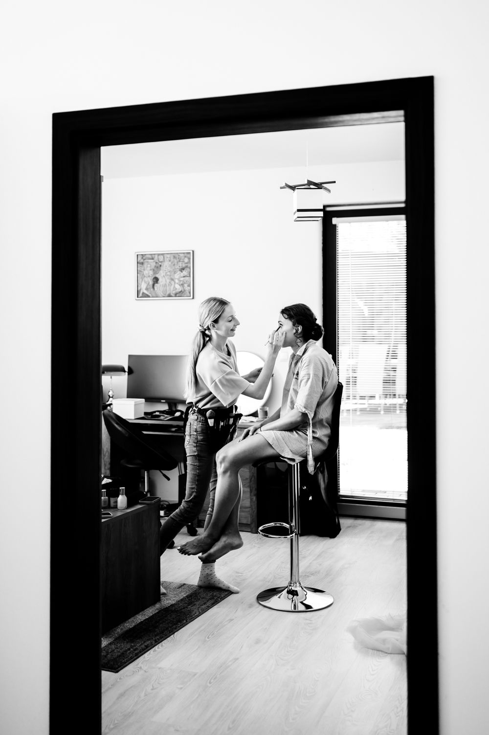 Black and white photo of a makeup artist applying makeup to a client seated on a bar stool in a modern room.