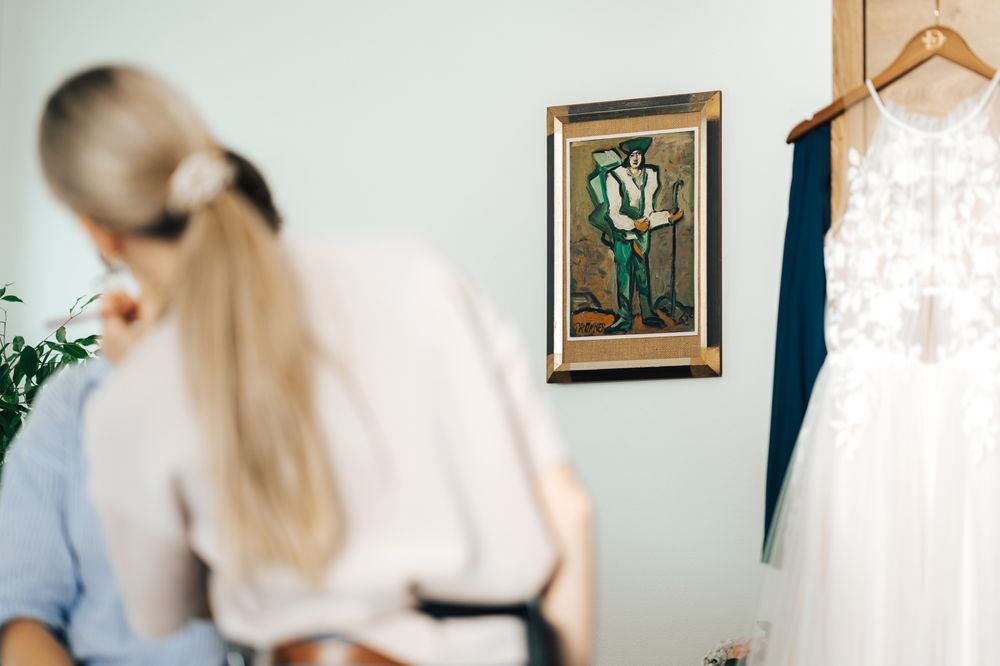Bride getting ready with lace wedding dress hanging near framed artwork in elegant bridal preparation room.