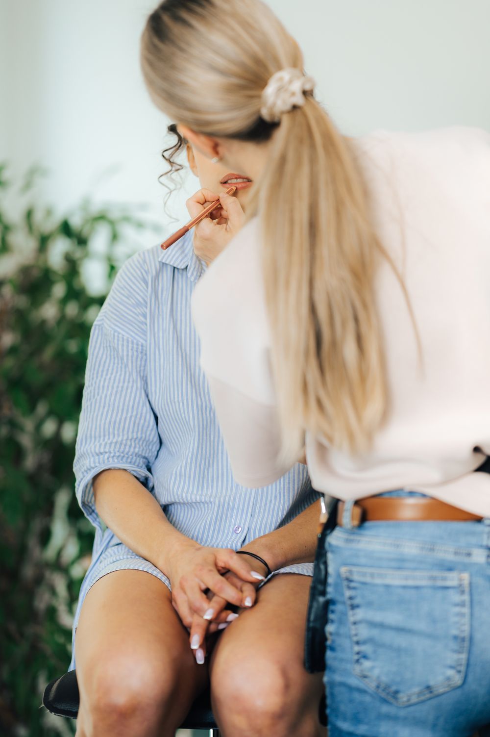 Makeup artist applying cosmetics to a seated blonde woman in a blue striped shirt in a bright studio.