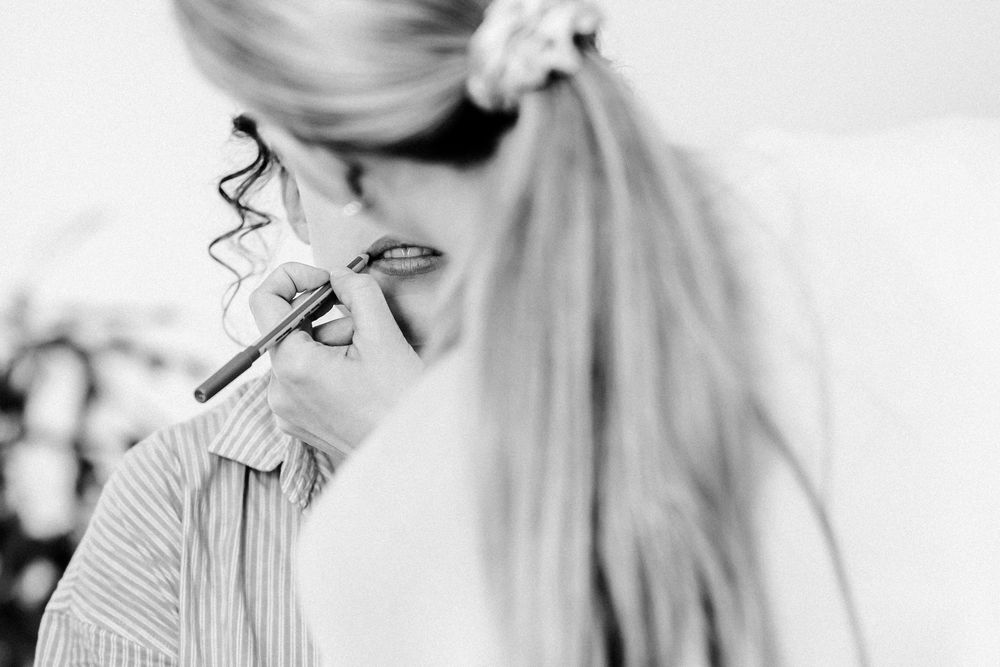 Black and white photo of a blonde woman applying makeup with a brush in a softly lit setting.