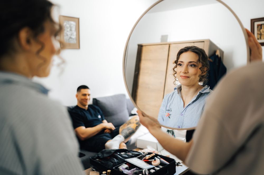 Woman applying makeup at mirror with man sitting on couch reflected in background during wedding preparation.